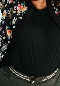 Persoon draagt een zwart shirt, een jas met een bloemenpatroon en groene broek met een zwart-wit gestreepte riem.