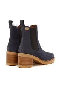 Botas de tobillo de ante azul oscuro con paneles elásticos en los laterales y un tacón de madera apilado. Suela texturizada notable y etiqueta de marca en la parte posterior.