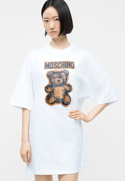 Νεαρή γυναίκα με κοντά μαύρα μαλλιά φοράει υπερμεγέθη λευκό φόρεμα με μεγάλο καφέ και μαύρο αρκουδάκι και το κείμενο "MOSCHINO" στο εμπρός μέρος.