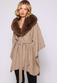 Femme blonde portant une cape beige ceinturée avec un grand col en fourrure marron sur un pantalon noir, se tenant devant un fond clair.