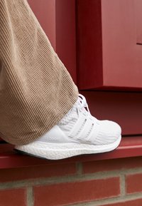 Fot i vit stickad Adidas Ultra Boost-sneaker som står på ett röd tegelsteg, iförd beiga kordflöjlsbyxor.
