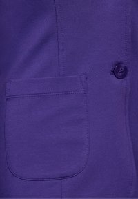 Gros plan sur un tissu violet avec une poche écusson cousue et un bouton violet assorti sur un vêtement.