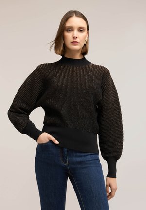 Žena v černém žebrovaném svetru s nafouklými rukávy a modrých džínách stojí před jednobarevným béžovým pozadím.