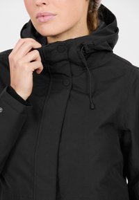 Svart vattentät jacka med hög krage, tryckknappar, justerbar dragsko på huvan och texturerat material. Närbild av framsidan.