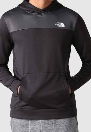 Person trägt einen schwarzen und dunkelgrauen North Face Hoodie mit den Händen in der Fronttasche, kombiniert mit hellgrauen Hosen.