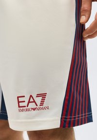 Κοντινό πλάνο σε λευκό σορτς με κόκκινες και ναυτικές κάθετες ρίγες στο πλάι και κόκκινο λογότυπο EA7 Emporio Armani κοντά στο τελείωμα, φορεμένο από ένα άτομο.