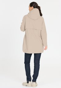 Veste imperméable beige avec capuche, dotée d'une coupe ample et d'une taille élastique. Portée par-dessus un pantalon en denim foncé et des bottines beiges.