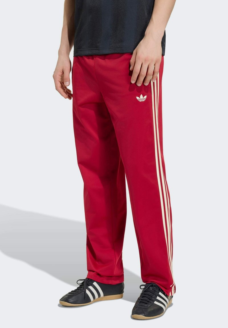 Mand iført røde Adidas træningsbukser med hvide sideløb og logo, kombineret med sorte Adidas sneakers med hvide striber.