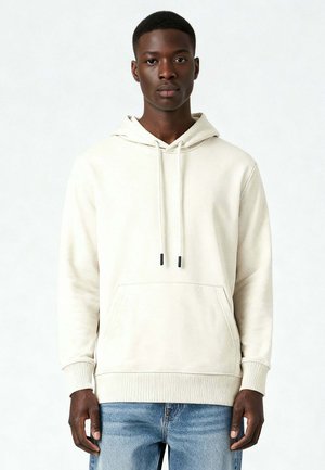 Jonge zwarte man staand, gekleed in een lichtbeige hoodie met trekkoorden en een blauwe spijkerbroek, kijkt recht voor zich uit tegen een effen achtergrond.