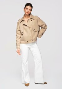 Femme portant un trench court beige, un pantalon large blanc et des chaussures marron pointues, posant devant un fond clair uni.