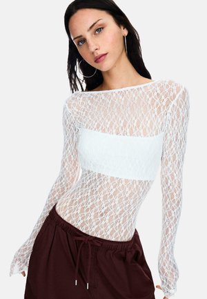 Witte kanten bodysuit met lange mouwen en een solide witte strook over de torso, gecombineerd met donker bordeauxrode shorts. Gedetailleerd kanten patroon zichtbaar.