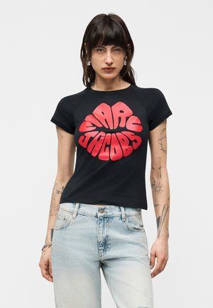 Donna con capelli scuri che indossa una t-shirt nera Marc Jacobs con testo in rosso audace e jeans azzurri, in piedi davanti a uno sfondo semplice.