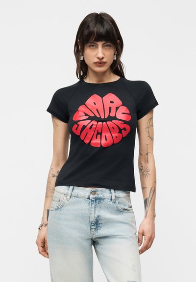 Femme aux cheveux foncés portant un t-shirt noir Marc Jacobs avec un texte rouge vif et un jean bleu clair, debout devant un fond uni.