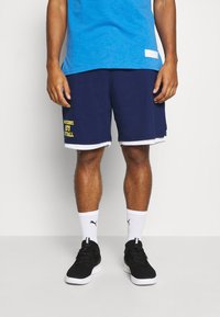 Man som bär blå sportshorts med gul text, vita Puma-strumpor, svarta sneakers och en blå T-shirt, stående framför en neutral bakgrund.