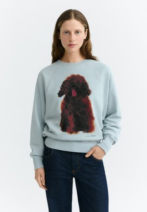 Junge Frau mit langen braunen Haaren trägt ein hellblaues Sweatshirt mit einer großen schwarzen und braunen flauschigen Hundeillustration, kombiniert mit dunklen Jeans.