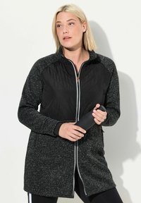 Schwarze und graue Zip-Jacke mit einer gesteppten Vorderseite und strukturierten Ärmeln, mit einem hohen Kragen und Seitentaschen; kombiniert mit einer schwarzen Wasserflasche.