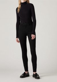 Femme portant un pull à col roulé noir côtelé, un jean skinny noir taille haute et des mocassins noirs sur un fond clair uni.
