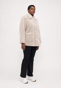 Beige, teddy-style coat with a collar and side pockets, featuring a soft, textured fabric. Paired with black pants and white sneakers.