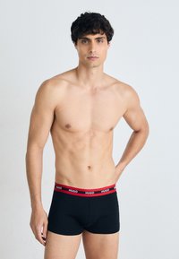 Schwarze Boxershorts mit einem roten elastischen Bund, der die Marke "HUGO" zeigt. Glatte Textur, enganliegendes Design und klassischer Stil.