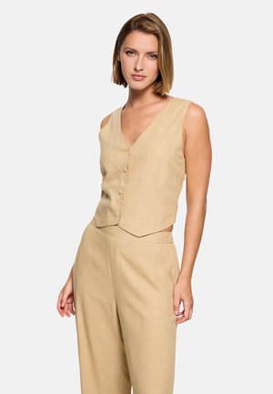 Mujer con chaleco beige sin mangas abotonado y pantalones de talle alto y pierna ancha a juego, frente a un fondo blanco.