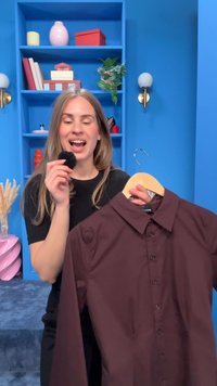 Γυναίκα κρατά ένα καφέ πουκάμισο με κουμπιά σε μια κρεμάστρα στο ένα χέρι και ένα μικρό μαύρο χνουδωτό αντικείμενο κοντά στο στόμα της σε ένα μπλε δωμάτιο με ράφια και φυτά.