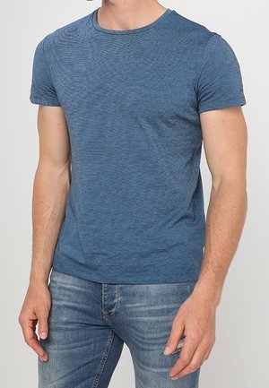 A man wearing a fitted short-sleeve blue crew neck t-shirt and light blue jeans standing against a plain background.