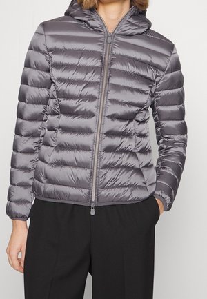 Personne portant une doudoune matelassée grise brillante avec capuche, fermée par une fermeture éclair, associée à un pantalon noir, une main dans la poche.