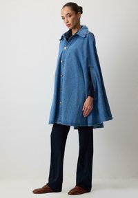 Denim kappe med en struktureret krave, gyldne knapper og sideåbninger. Bæret over mørke bukser og lædersko. Glat tekstur, levende blå farve.