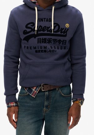 Mand iført en navy hoodie med teksten "SuperDry", ternet skjorte under, blå jeans, brun bælte og et guldkronometer, stående mod en hvid baggrund.