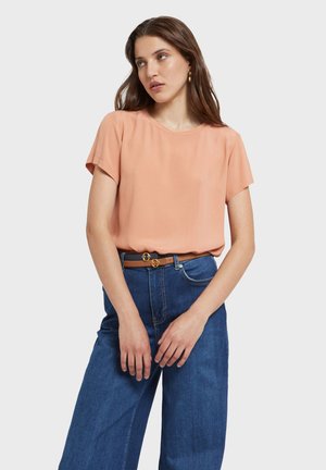 Jonge vrouw in een perzikkleurige blouse met korte mouwen, ingestopt in een blauw spijkerbroek met hoge taille en een bruine riem, staand tegen een effen achtergrond.