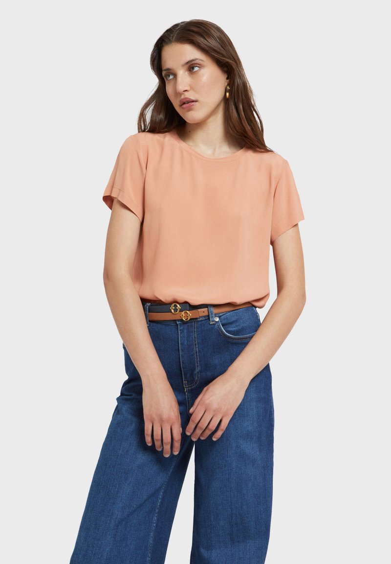 Jeune femme portant un chemisier pêche à manches courtes rentré dans un jean bleu taille haute avec une ceinture marron, debout devant un fond uni.
