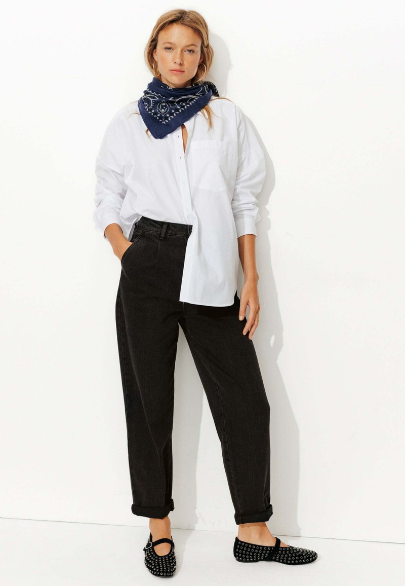 Chemise blanche à boutons, pantalon noir taille haute, écharpe marine à motifs et ballerines noires avec des accents cloutés. Tissu lisse et coupe décontractée.
