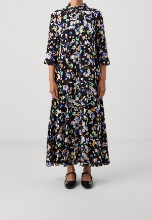 Kvinde iført en sort, lang blomsterkjole med krave og trekvartærmer, kombineret med sorte Mary Jane-sko.