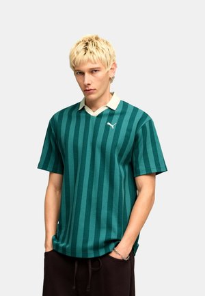 Uomo biondo che indossa una polo a maniche corte verde a righe con colletto bianco, pantaloni neri e un braccialetto a catena al polso sinistro.