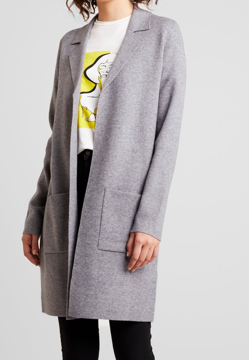 Manteau en laine grise à coupe décontractée, manches longues et deux poches avant. Il présente un col cranté et une texture lisse.