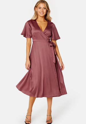 Klänning i mörk mauve, tillverkad av mjukt satin. Har korta volangärmar, en djup V-ringning och ett knytbälte i midjan. Längden når till vaden.