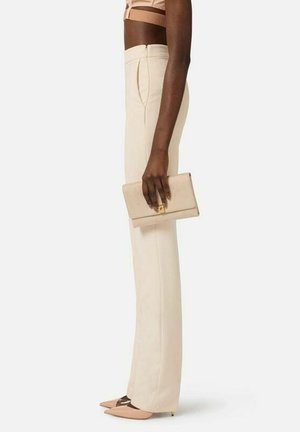 Kvinde iført beige bukser med høj talje, nøgen spids hæl, holder en matchende beige clutch, stående sideværts mod en ensfarvet baggrund.