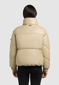 Beige pufi dzseki magas gallérral, steppelt kialakítással és rugalmas mandzsettákkal. Simá, fényes felület; hátul kis logó található.