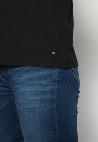 Camiseta de algodón negra con un pequeño logo rojo, blanco y azul en el dobladillo, combinada con unos vaqueros de mezclilla azul con una superficie texturizada.