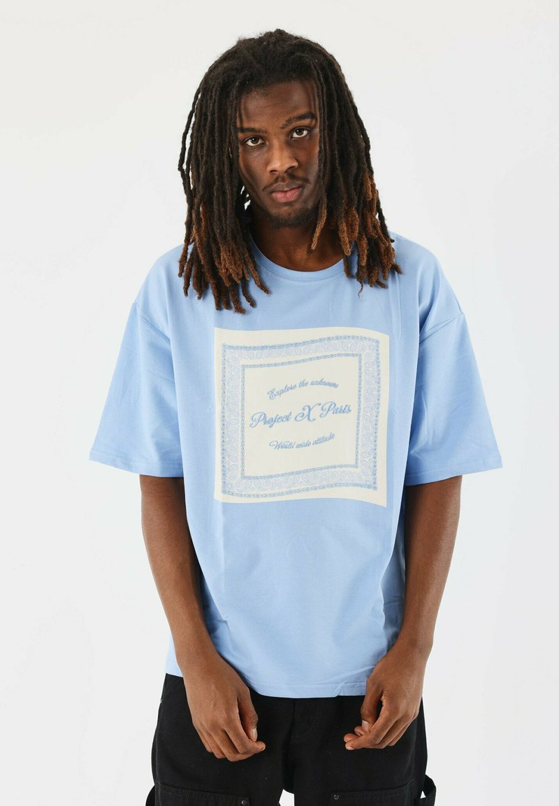Camiseta de corte oversized en azul claro, hecha de algodón, con un diseño gráfico central en texto blanco y azul dentro de un borde decorativo.