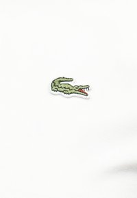 Πράσινο κεντημένο λογότυπο κροκόδειλου με ανοιχτό στόμα, λεπτομερή δόντια και κόκκινη γλώσσα σε λευκό υφασμάτινο φόντο.