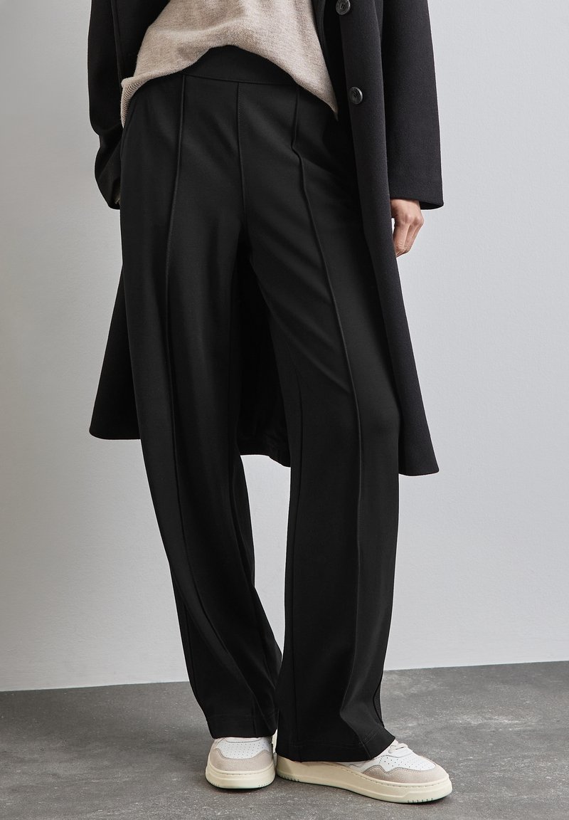 Personne portant un pantalon noir à jambes larges, un pull beige, un long manteau noir et des baskets blanches, debout sur un sol gris devant un mur clair.