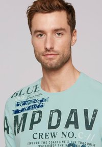 Jasnoniebieska koszulka z krótkim rękawem z wyraźnym czarnym tekstem i niebieskimi akcentami graficznymi. Materiał wydaje się gładki i lekki.