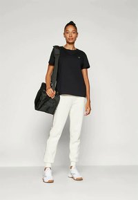 Juodas marškinėlis, balti sportiniai džinsai ir balti sportbačiai. Modelis laiko juodą maišą. Lygus audinys, laisvas siluetas, subtilus prekės ženklo ženklinimas ant marškinėlių.