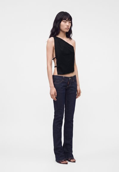Femme aux longs cheveux noirs portant un top noir asymétrique avec des découpes latérales et un jean bleu foncé droit avec des sandales, se tenant face à l'avant.