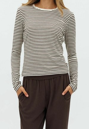 Vrouw die een wit shirt met lange mouwen draagt met dunne zwarte horizontale strepen en losse donkerbruine broek met handen in de zakken.