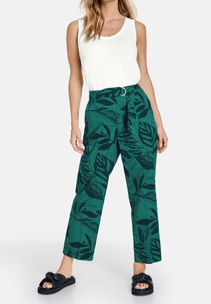Femme portant un top blanc sans manches, un pantalon vert taille haute à jambes larges avec un imprimé de feuilles foncé et des sandales noires à enfiler.