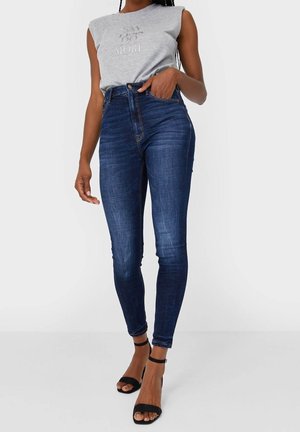 Frau trägt dunkelblaue High-Waist Skinny-Jeans, graues ärmelloses Oberteil mit dem Schriftzug "SAY LESS DO MORE" und schwarze Riemchen-Absatzschuhe, steht mit der Hand in der Hosentasche.
