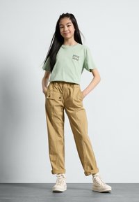 Mintgrønn T-skjorte med en liten grafisk lomme, kombinert med beige cargo-bukser med elastisk midje og hvite høyhælte sneakers.
