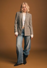 Blazer a quadri con due pattine frontali, top color crema e jeans blu a gamba larga. Stivaletti marroni completano l'outfit su uno sfondo sobrio.